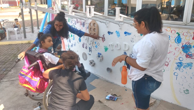 fotografia de 4 pessoas, incluindo uma cadeirante, na frente de um muro tátil, no qual é possível perceber diversos objetos fixados e pendurados