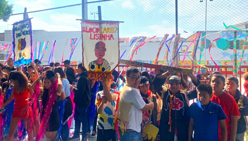 Estudantes fantasiados e com estandartes do blocos de carnaval na quadra da escola.