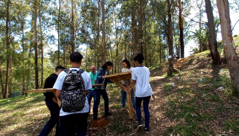 Fotografia de estudantes e professor em área verde carregando mesas de madeira.