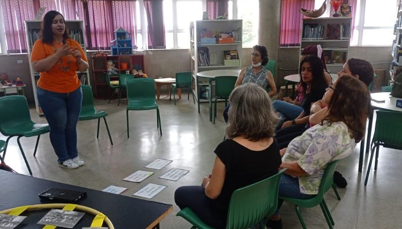 Pessoas estão sentadas em roda enquanto uma mulher, que está de pé no centro, conversa com elas.