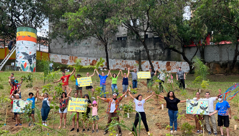 Pessoas estão de pé em área verde com os braços levantados e as mãos dadas. Algumas delas seguram cartazes com desenhos de pássaros e escritas "Sejam bem-vindos ao bosque". Entre eles há mudas de árvores de tamanho médio.