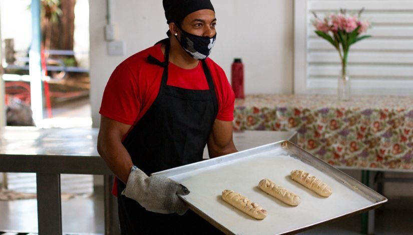 Homem de camiseta vermelha e avental preto usando luvas culinárias e segurando uma forma untada com três pães trançados.