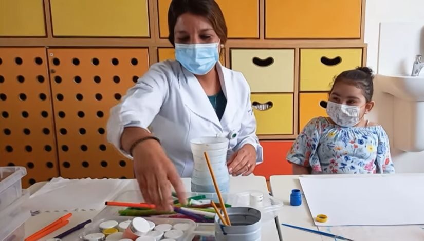 Fotografia de uma professora e uma criança sentadas a mesa com materiais para pintura como pincéis, tintas, folhas brancas de papel, entre outros.