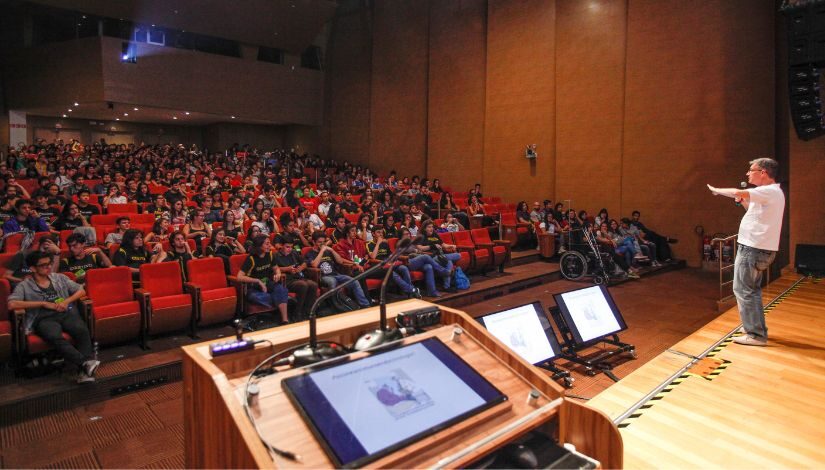 Fotografia de um auditório com inúmeras pessoas sentadas nas cadeiras e um homem no palco falando com elas.