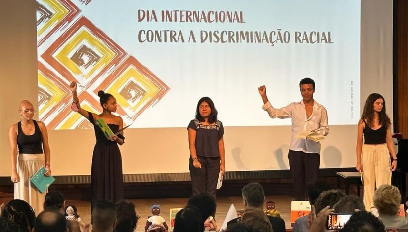 Foto de 5 estudantes no palco, ao fundo, uma projeção com os dizeres Dia Internacional contra a discriminação racial