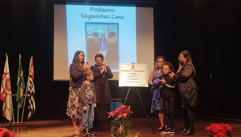 Fotografia de quatro mulheres adultas e duas crianças em um palco com uma placa ao centro. Ao fundo projetado em uma tela a fotografia de uma mulher escrito Professora Nazaré Neri Lima.