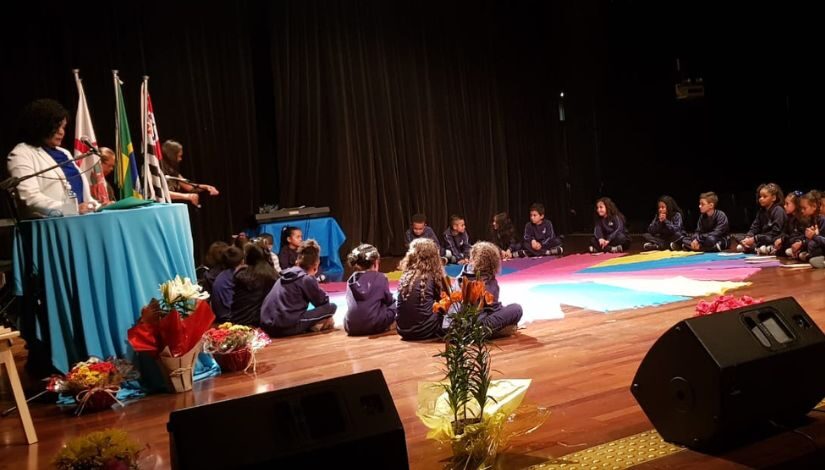 Fotografia de crianças sentadas em círculo em um palco. Ao centro delas no chão um pano circular multicolorido.