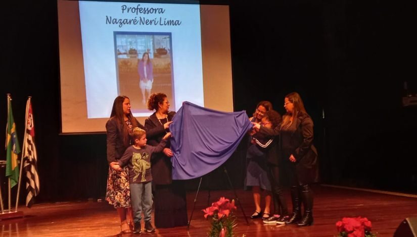 Fotografia de quatro mulheres adultas e duas crianças no palco descerrando a placa ao centro. Ao fundo projetado em uma tela a fotografia de uma mulher escrito Professora Nazaré Neri Lima.