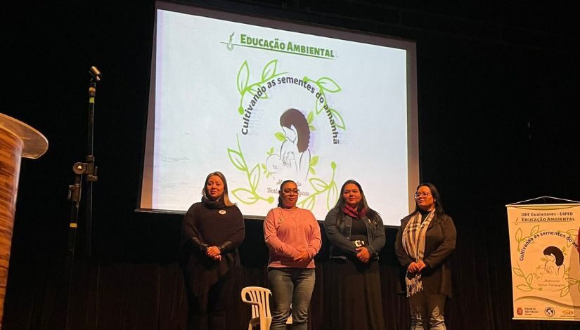 Fotografia de quatro mulheres no palco. Ao fundo, um telão escrito Educação Ambiental.