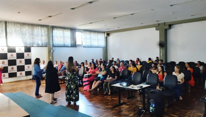 Um auditório com um grupo de 40 pessoas sentadas assistindo uma apresentação sendo feita por 3 pessoas que estão em pé.