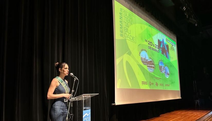 Imagem mostra palco com telão e imagem projetada onde se lê "Semana da Sustentabilidade". Jovem com uma faixa fala ao púlpito.