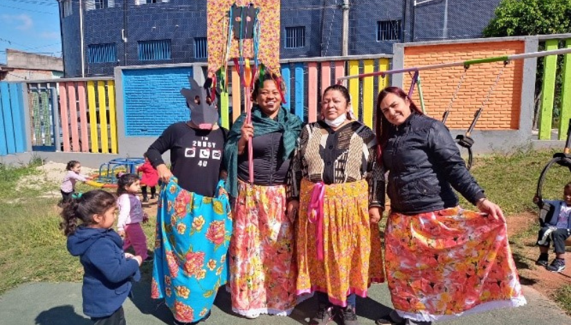 Quatro mulheres estão de pé posando para foto, elas estão usando saias rodadas com estampas floridas. A mulher da ponta esquerda está usando uma máscara de boi, a que está ao seu lado segura um estandarte com fitas coloridas e o desenho do rosto de um boi. 