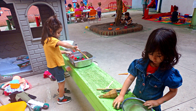 Duas meninas estão brincando de fazer comida, uma delas segura uma colher e mexe tampinhas de garrafa que estão em um pote, e a outra segura uma peneira. Elas estão apoiadas em um banco verde, no chão, atrás delas, há algumas bonecas. Atrás delas pode-se ver outras crianças sentadas no chão, uma árvore, uma casinha e ainda outras crianças sentadas em cadeiras em volta de uma mesa.