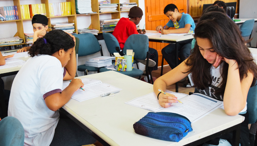 Fotografia de sala de aula mostra estudantes sentados em suas carteiras escolares em duplas, um de frente pro outro. Eles estão escrevendo em folhas que estão em cima das mesas.