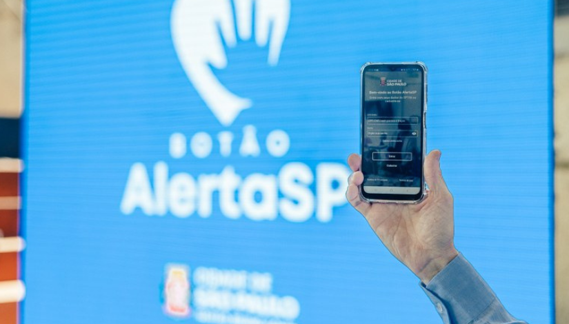 Fotografia de uma mão segurando um celular. Ao fundo há um painel na cor azul, no centro há um o desenho de um coração em branco e de uma mão em azul com o texto "Botão AlertaSP" .  Abaixo há a logomarca da cidade.