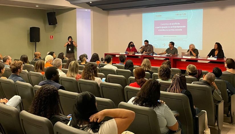 Imagem de pessoas sentada em um auditório, à mesa de apresentação há cinco pessoas e ao uma imagem projetada.