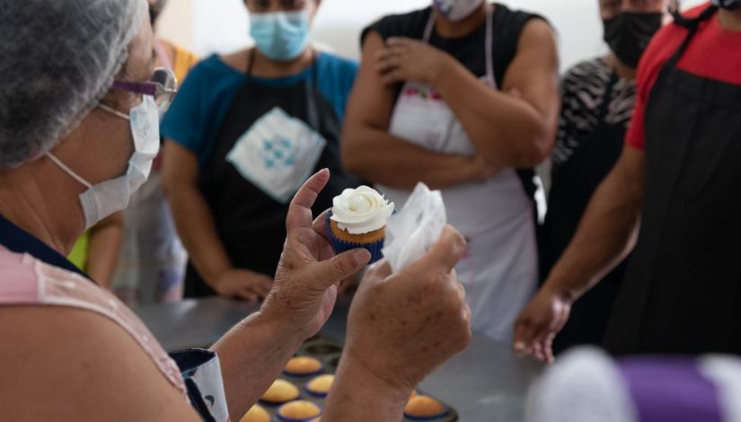 Fotografia de uma professora no curso de confeitaria do CMCT. Ela usa máscara, toca e segura um cupcake com topo de chantilly.