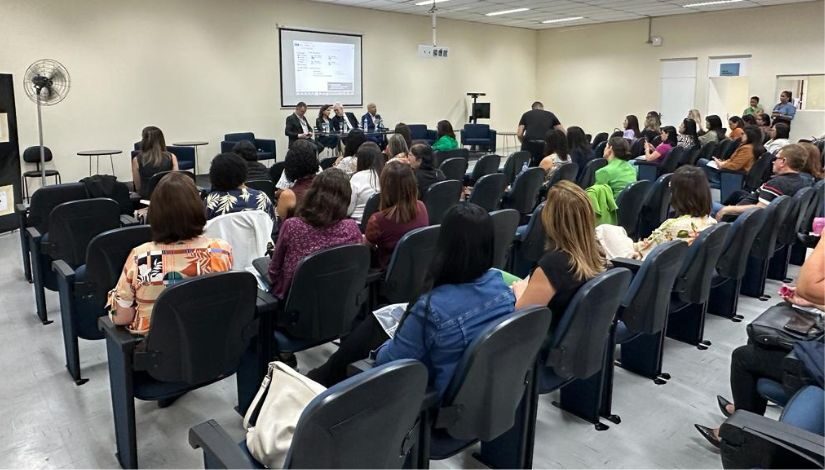 Plateia de pessoas sentadas em suas cadeiras, elas estão de costas para a fotografia e ao fundo um projetor e quatro pessoas sentadas a mesa.