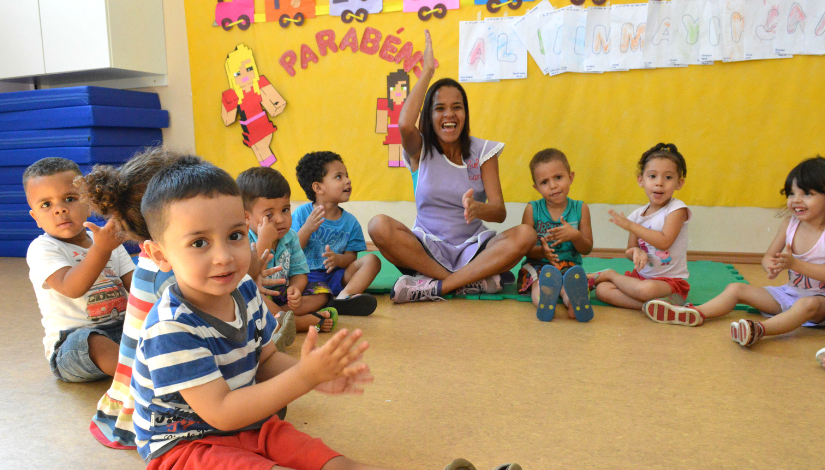 Fotografia mostra várias crianças sentadas em roda no chão. Entre elas está uma professora, ela usa jaleco lilás e está batendo palmas.