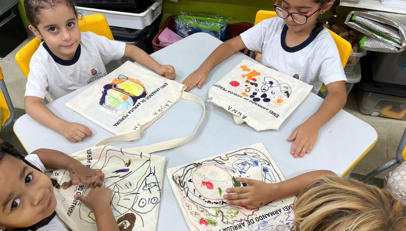 Crianças pequenas sentadas às mesa com sacolinhas de leitura onde se lê o nome da escola "EMEI Armando Arruda Pereira" e há também o desenho feito por elas.