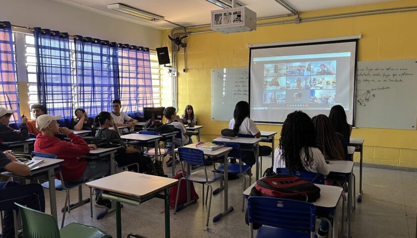 Sala de aula onde os estudantes estão sentados de costas para a foto. Na frente deles há uma tela mostrando uma vídeo chamada. Eles veem diversos quadrados pequenos com as pessoas que estão participando da ligação.