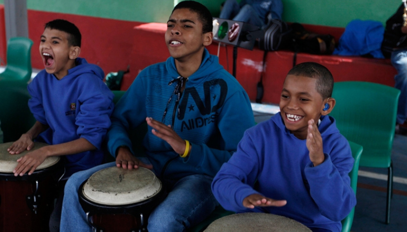 Três meninos estão sentados. Entre as pernas deles há o instrumento musical Timba. Os meninos batem nos instrumentos com as mãos.