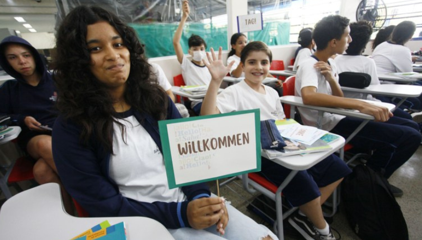 Menina está segurando uma placa onde está escrito "willkommen", ela está numa sala de aula e ao fundo pode-se ver outros estudantes.