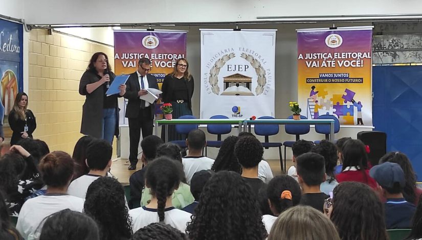 Fotografia de um auditório com crianças sentadas viradas para o palco. no palco três adultos, uma mesa com cinco cadeiras e três banners sobre justiça eleitoral.