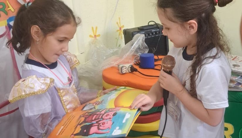 Fotografia de duas crianças: uma menina está fantasiada e segura o livro de literatura infantil com a imagem de três porquinhos e a outra usa uniforme escolar e segura um microfone.