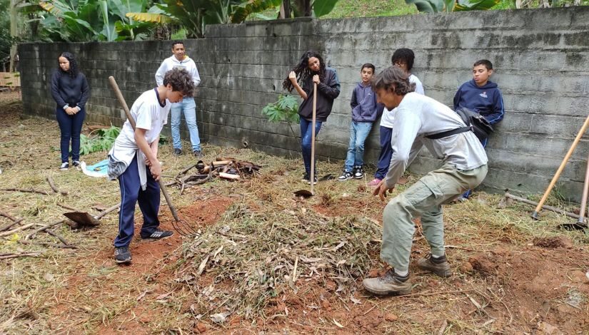 Grupo de estudantes e um adulto estão em área externa com enxadas e outros itens para manusearem a terra. No centro da imagem o adulto e um estudante estão ao redor de resíduos de folhas e terra.