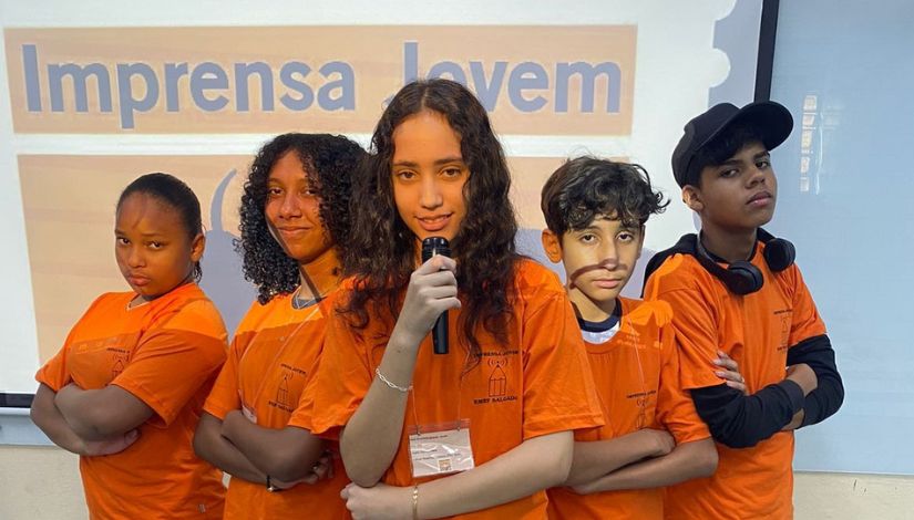 Cinco estudantes com camiseta laranja do Projeto Imprensa Jovem posam para fotografia, a estudante que está ao centro segura um microfone. 