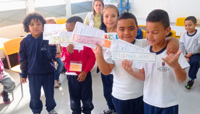 Fotografia crianças segurando plaquinhas com seus próprios nomes, elas estão sorrindo para a foto. Pode-se ver plaquinhas com os nomes “Davi Vinicius” e “Pedro Lucas”