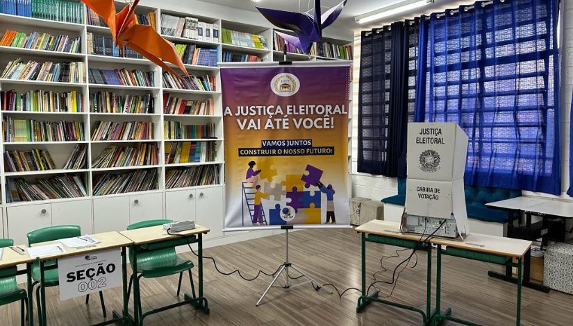 Sala com estantes de livros ao fundo preparada para a votação do Grêmio Estudantil com urna eletrônica da Justiça Eleitoral.