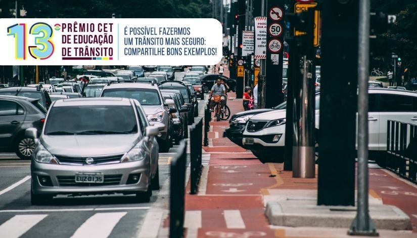 Fotografia mostra uma avenida com carros, faixa de pedestre, ciclovia, placas e ciclistas. No canto superior esquerdo está escrito 