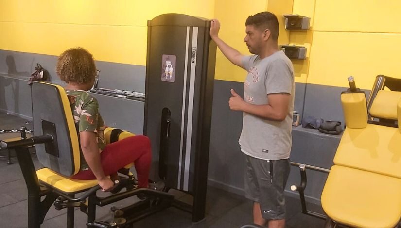 Mulher está sentada em equipamento de academia, um homem está de pé ao seu lado.