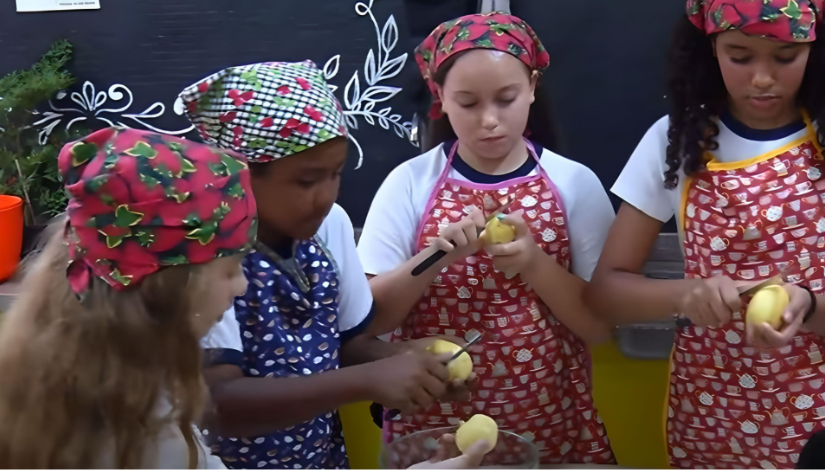 Quatro meninas estão em volta de uma mesa cortando batatas. Elas usam avental e touca estampada.