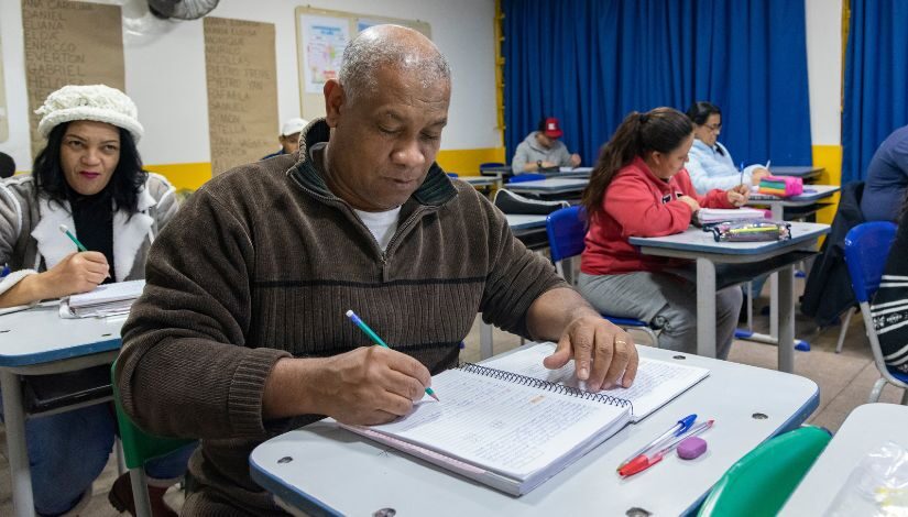 Fotografia de uma sala de aula da Educação de Jovens e Adultos. Em primeiro plano um homem negro vestindo blusa marrom está sentado a frente de sua classe e escreve com um lápis em seu caderno. Em segundo plano há outros cinco educandos, sendo três mulheres e dois homens.