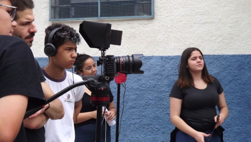 Estudantes e professor durante a gravação de um filme curta-metragem. Uma menina segura uma claquete de cinema e um aluno está atrás de um tripé com uma câmera. Outras três pessoas observam algo que está fora da captação pela fotografia. 