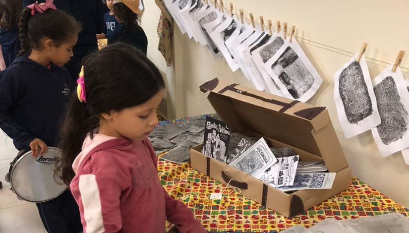 Fotografia de crianças de pé na frente de uma mesa, que está coberta com uma toalha florida. Sobre ela, há caixas com livros de cordel.