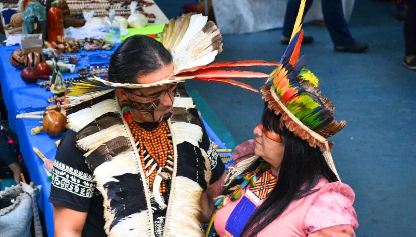 Fotografia mostra em primeiro plano uma mulher e um homem indígenas, os dois usam cocores de penas coloridas e estão se olhando. Ao fundo, elementos como chocalhos, colares e outros itens da cultura indígena.