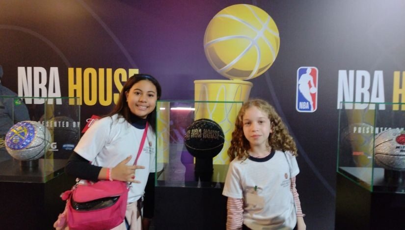 Fotografia de duas estudantes com o uniforme da prefeitura de SP posam para foto e ao fundo está escrito NBA House. Ao lado de cada uma há uma bola brilhante.