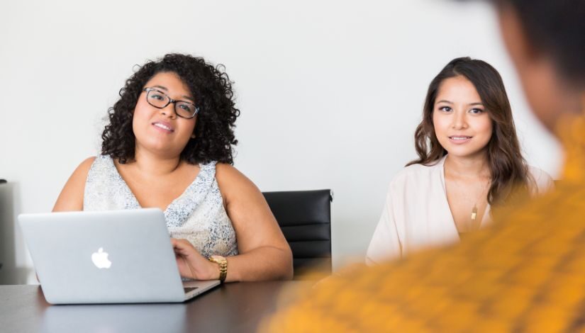 Duas mulheres sentadas a mesa viradas para a câmera, uma está com um computador portátil, e uma terceira pessoa que aparece de costas apenas a parte do ombro e da cabeça.