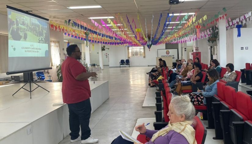 Fotografia de várias pessoas sentadas em cadeiras olhando para um homem que está a frente falando e atrás dele tem um telão com imagem projetada.