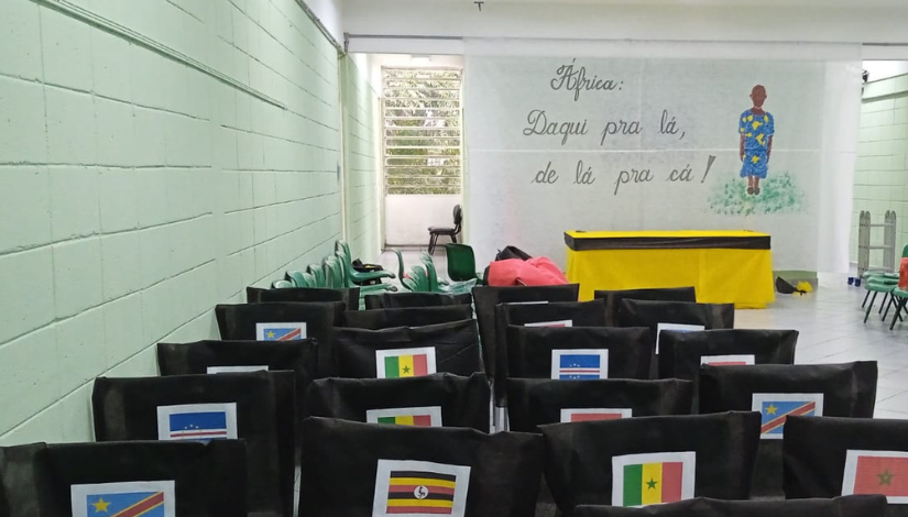 Fotografia de um espaço com cadeiras enfileiradas, elas estão com capas pretas e com bandeiras de países africanos colados. A frente há um painel com o desenho de uma pessoa negra e o texto "África: daqui pra lá, de lá pra cá!".