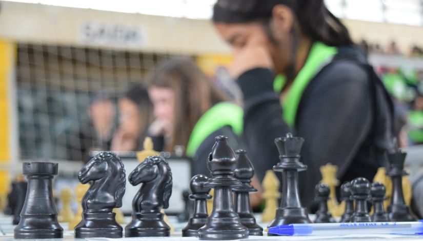 Fotografia com peças de xadrez pretas em primeiro plano; em segundo plano desfocado estudantes concentras em suas jogadas de xadrez.