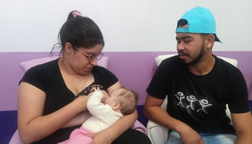 Fotografia de uma mãe amamentando sua bebê. Ela esta sentada em uma poltrona e ao seu lado está o seu companheiro que também olha para a bebê.