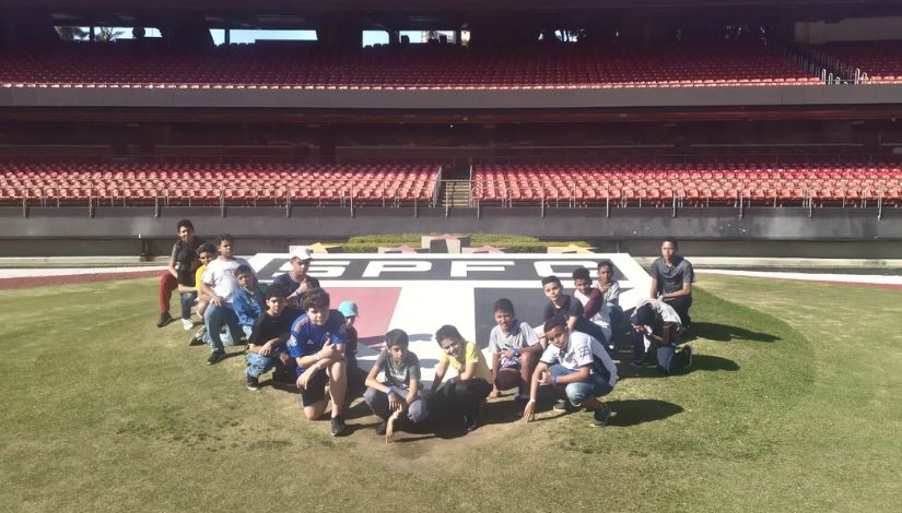Fotografia de crianças posando no gramado do estádio de futebol junto ao símbolo do São Paulo.