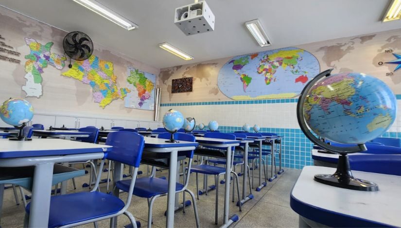 Fotografia de uma sala de aula com globos terrestres sobre a mesa e pintura nas paredes de mapas da cidade de São Paulo, do estado de SP, do Brasil e do mundo.