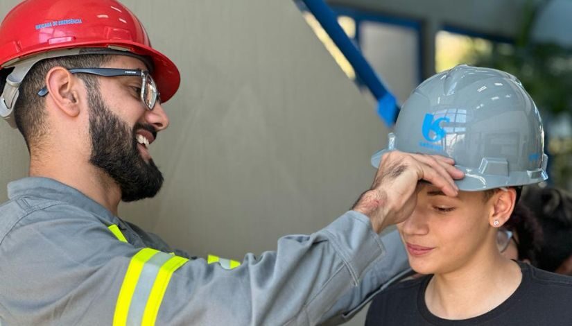 Fotografia de um homem com capacete vermelho e uniforme da Sabesp colocando um capacete cinza com a logomarca da Sabesp em um jovem.