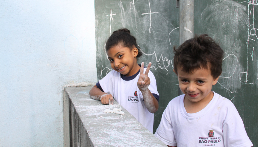 Fotografia de duas crianças. elas vestem uniforme escolar da prefeitura de São Paulo, olham para a câmera e sorriem 